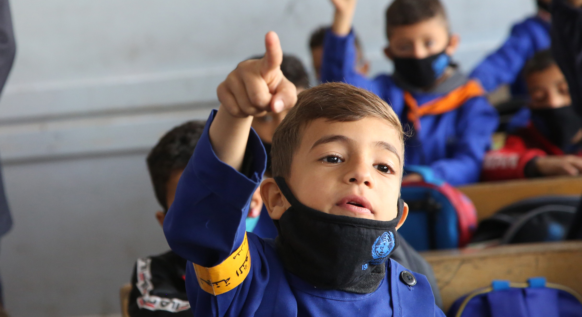 A boy raises his hand in a classroom. He is wearing a face mask with UNRWA's emblem.