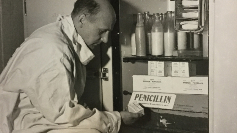 A man dressed in a white laboratory coat is looking at items inside a fridge which has boxes with a label that says Penicillin