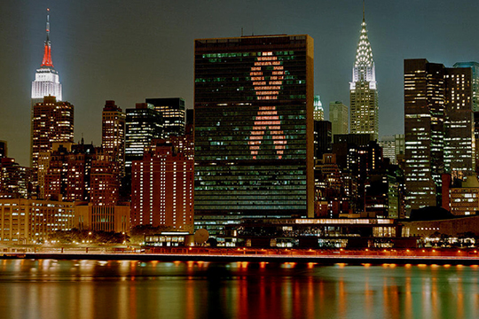 يُضاء مبنى الأمانة العامة للأمم المتحدة كشريط أحمر ويظهر النهر الشرقي في المقدمة.
