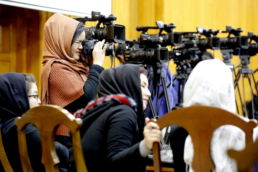A woman stands up holding a photo camera against a line-up of video cameras. 