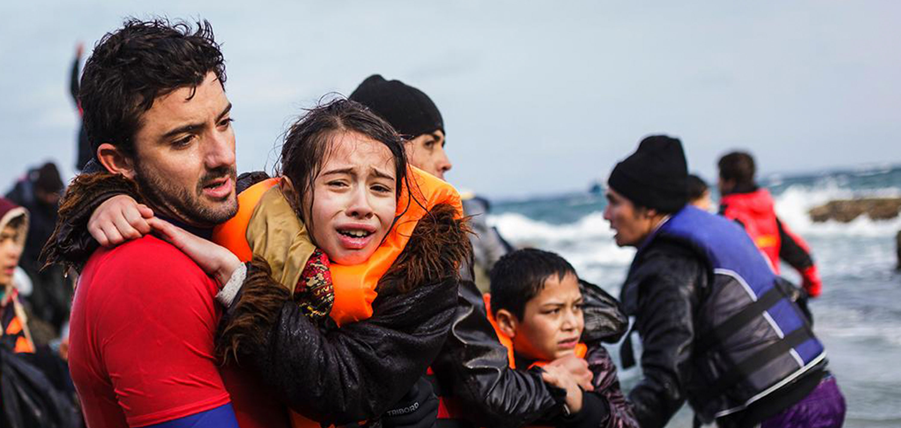 A man carries a girl-child wearing a safety vest. They are moving along watery shores amidst other refugees.