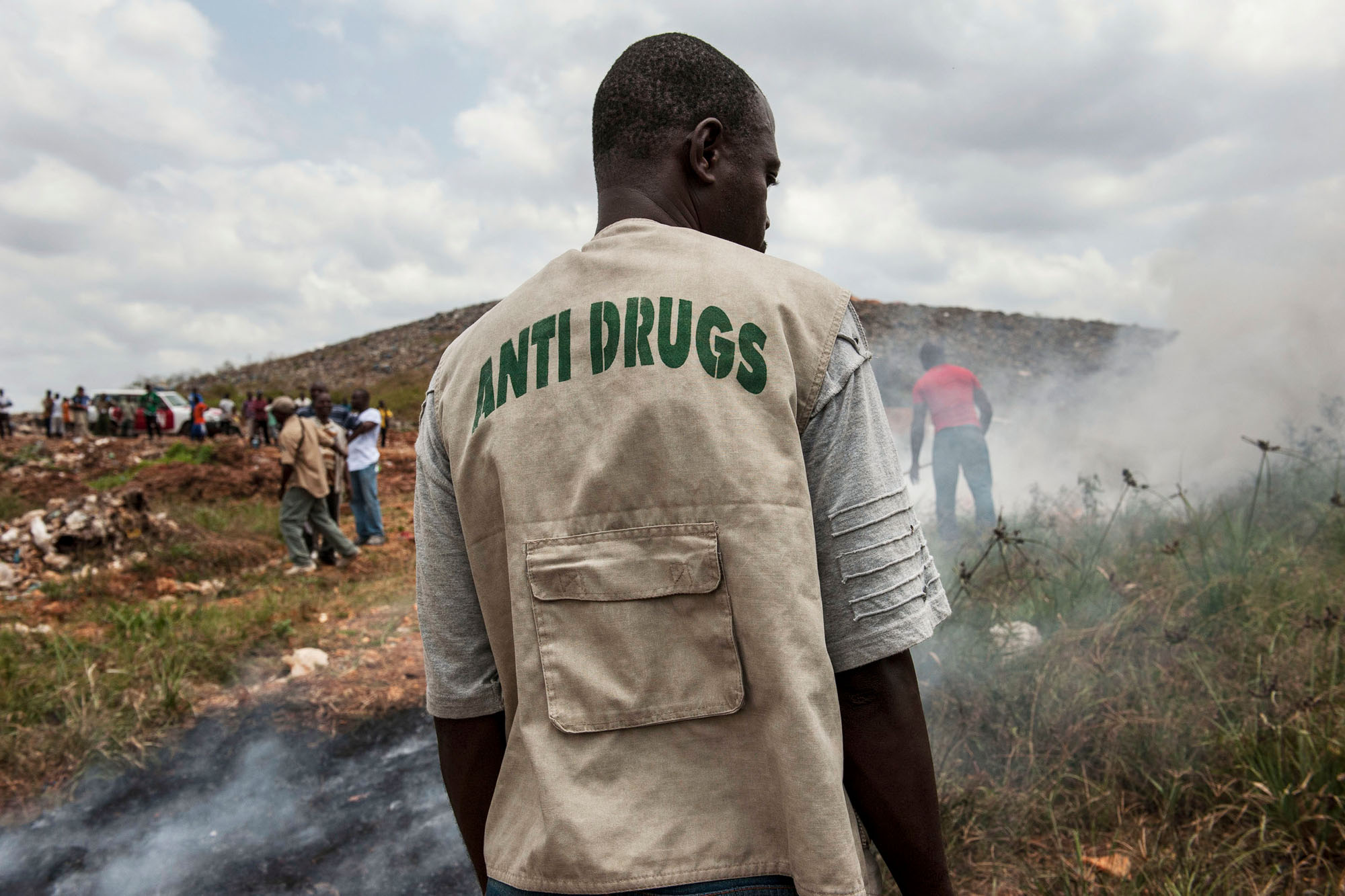 The back of a man wearing a vest that reads “Anti Drugs”, as he faces smoke.