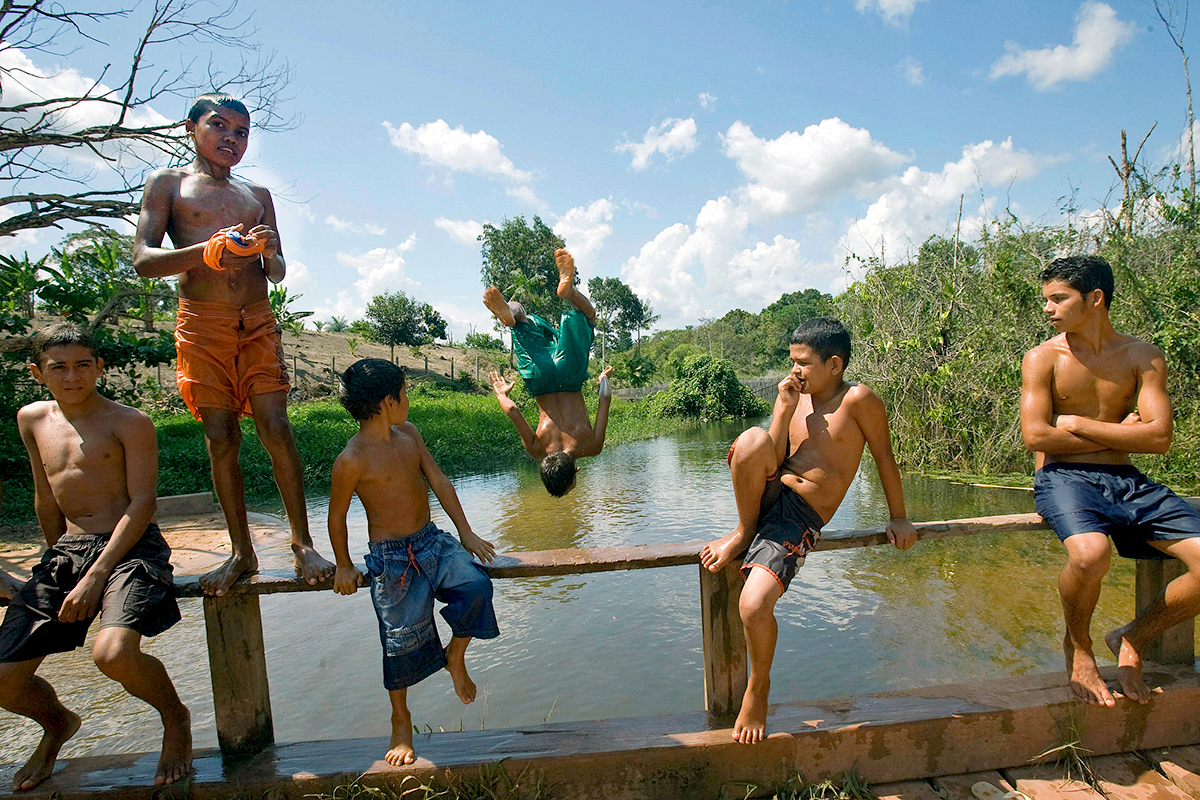 Boys sit on a barrier of the river while another is doing a mid-air flip into the river.  