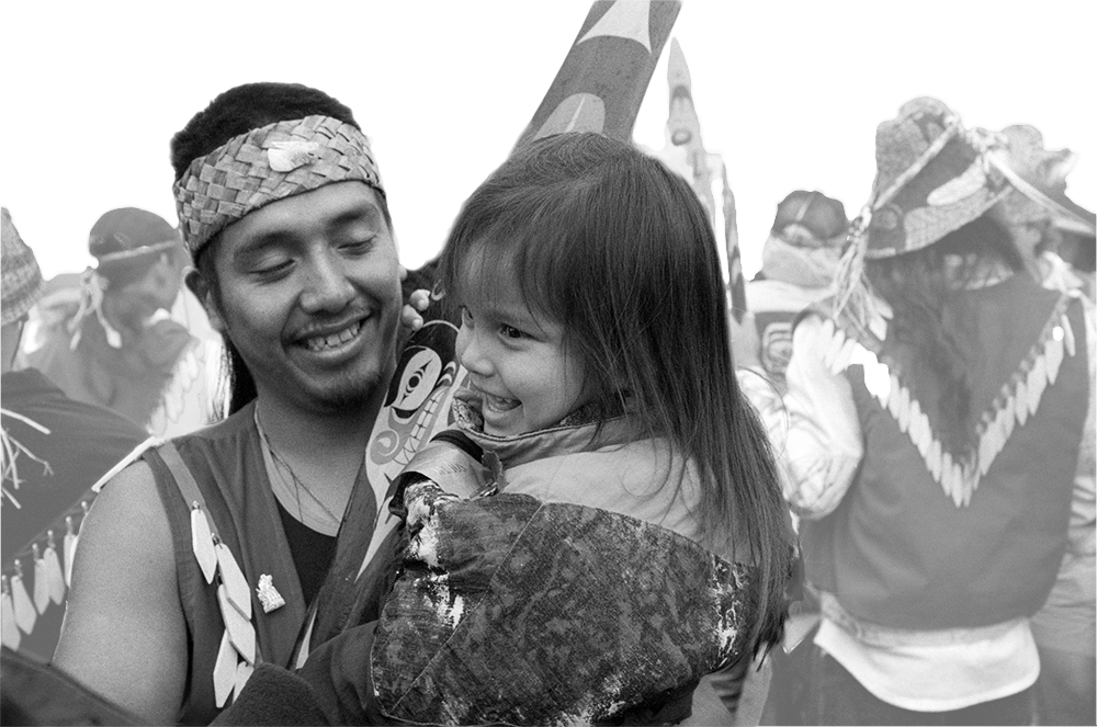 An indigenous father stands in a crowd holding his daughter and a paddle