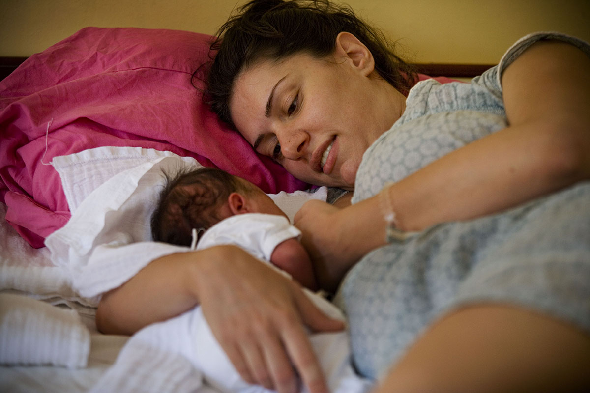 A woman lies in bed next to her newborn. 