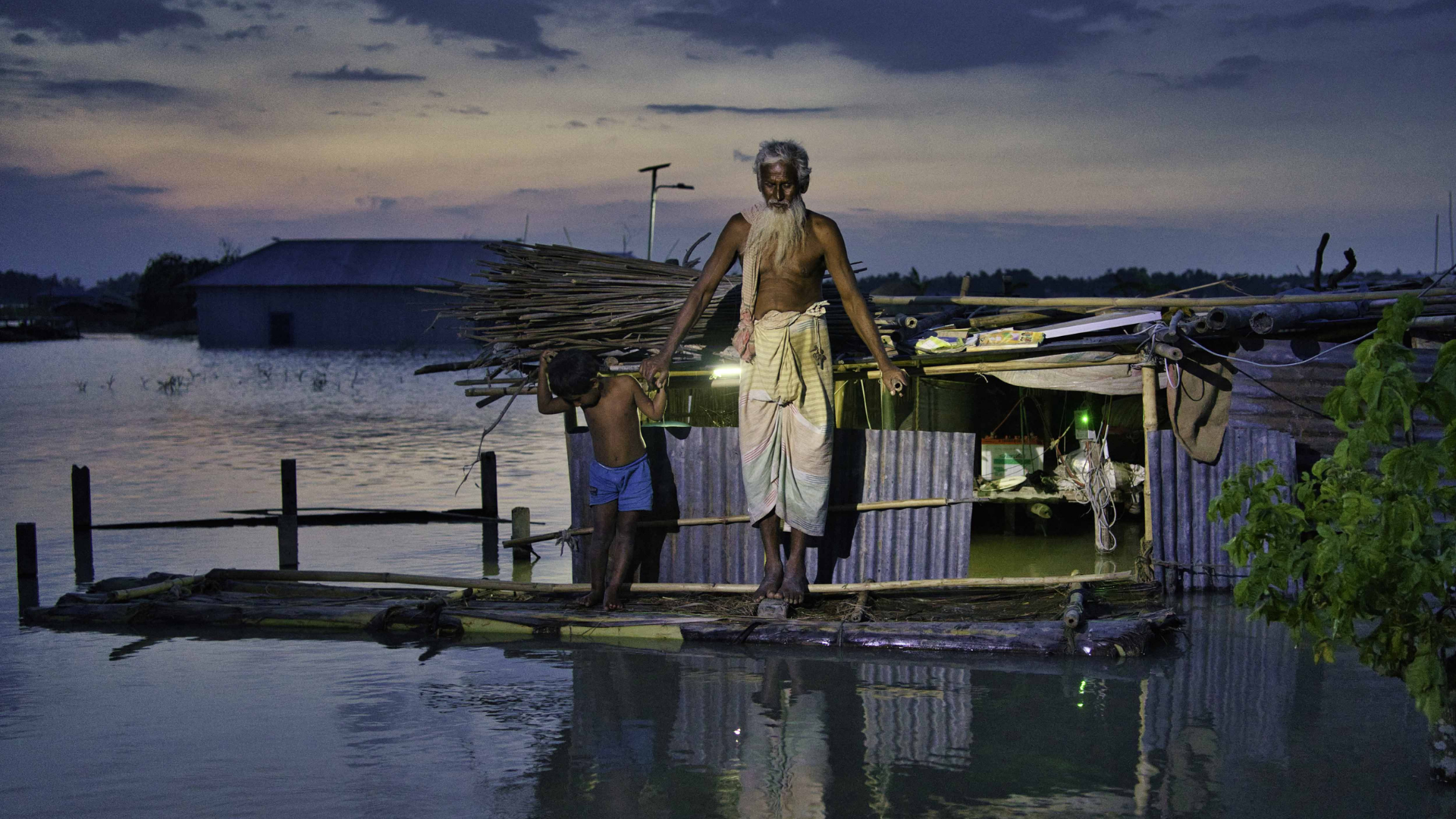 Abdus and his grandson stand on a raft made from banana trees in front of their submerged house in Char Bara Dhul, northern Bangladesh. The family sleep on a boat as they wait for the water to subside.