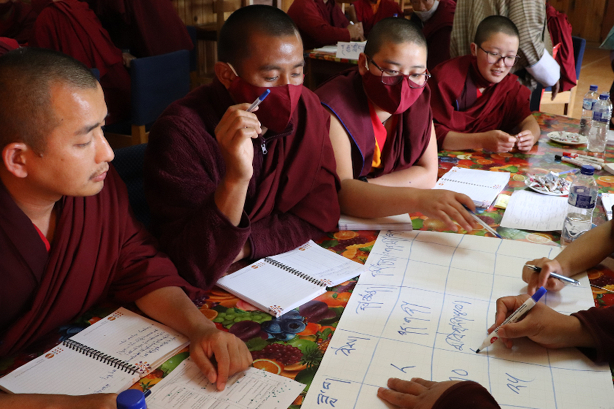 Monks wearing facemasks sit at a table with notebooks.