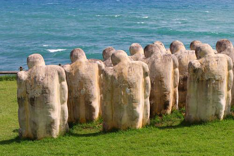 A set of sculptures depicting people with heads bowed and shoulders hunched, face the sea.