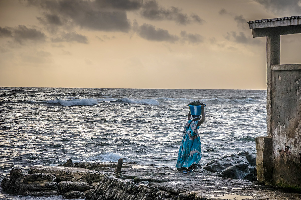 A woman carries a container on top of her head in front of a rough ocean