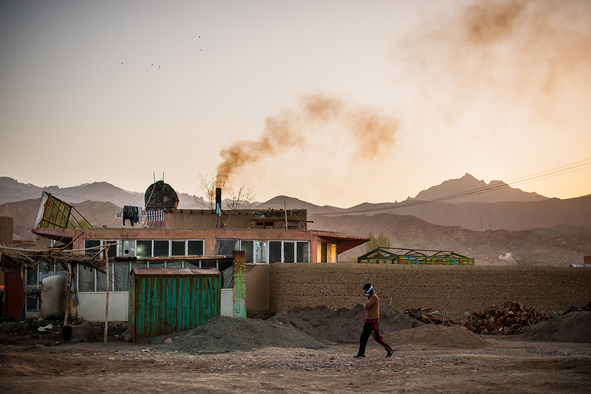 A man walks on a dirt path past a home in arid mountainous territory.