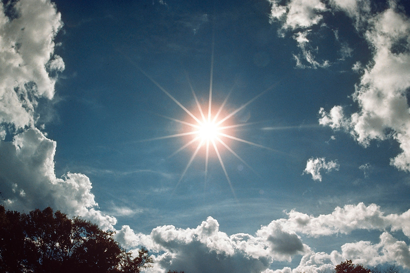 The sun shining brightly in a blue sky with clouds dispersed far apart around it.