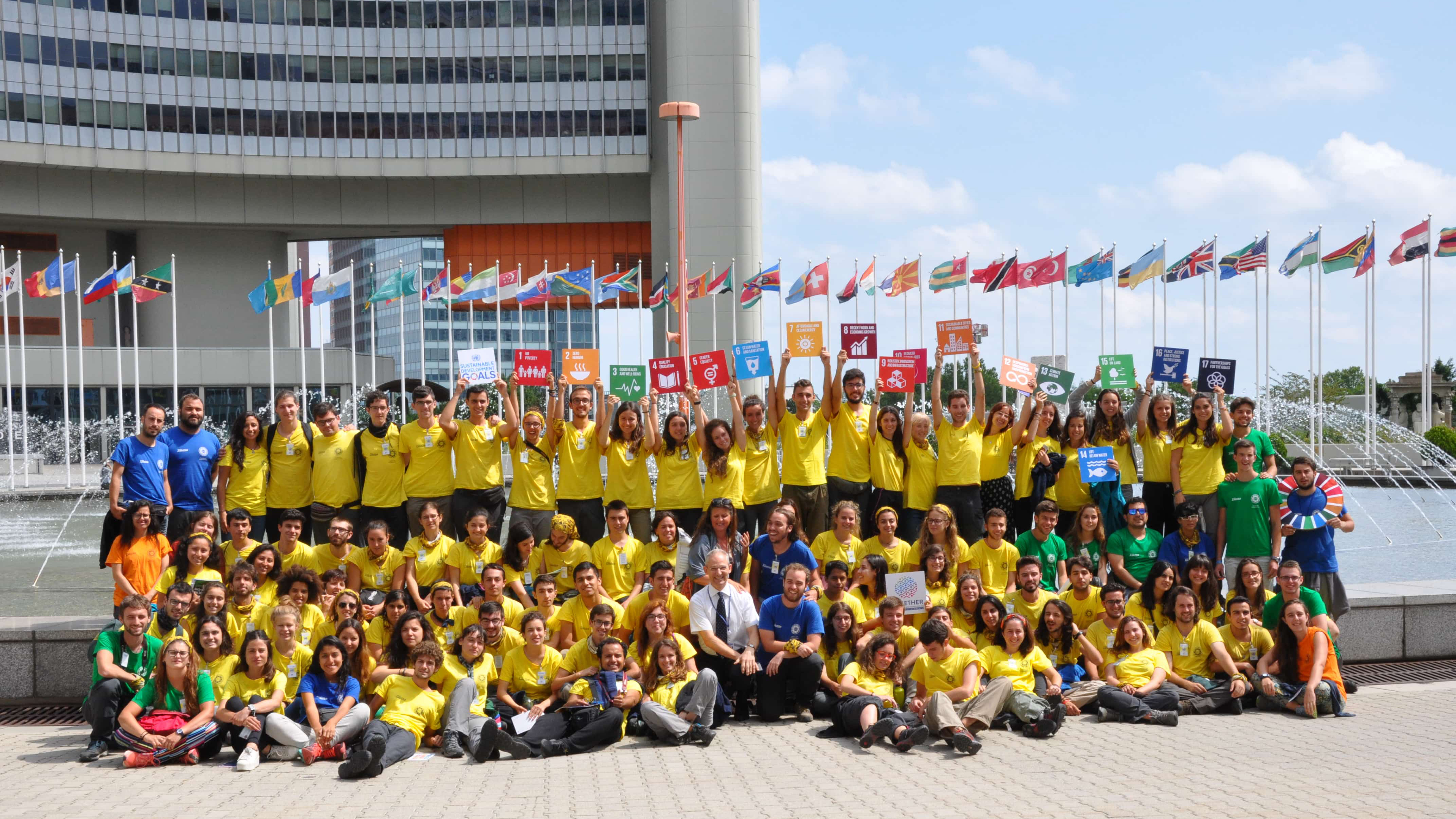A large group of children wearing yellow t-shirts hold up SDG signs outside the UN in Vienna