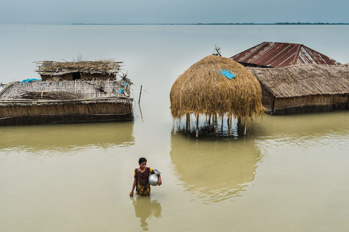 a woman carries a water container wading through water around flooded huts.