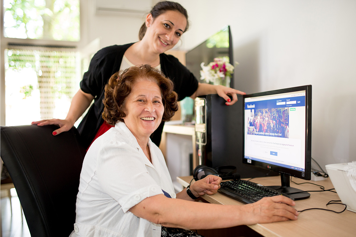 an older woman sits behind a computer with another woman standing behind her 