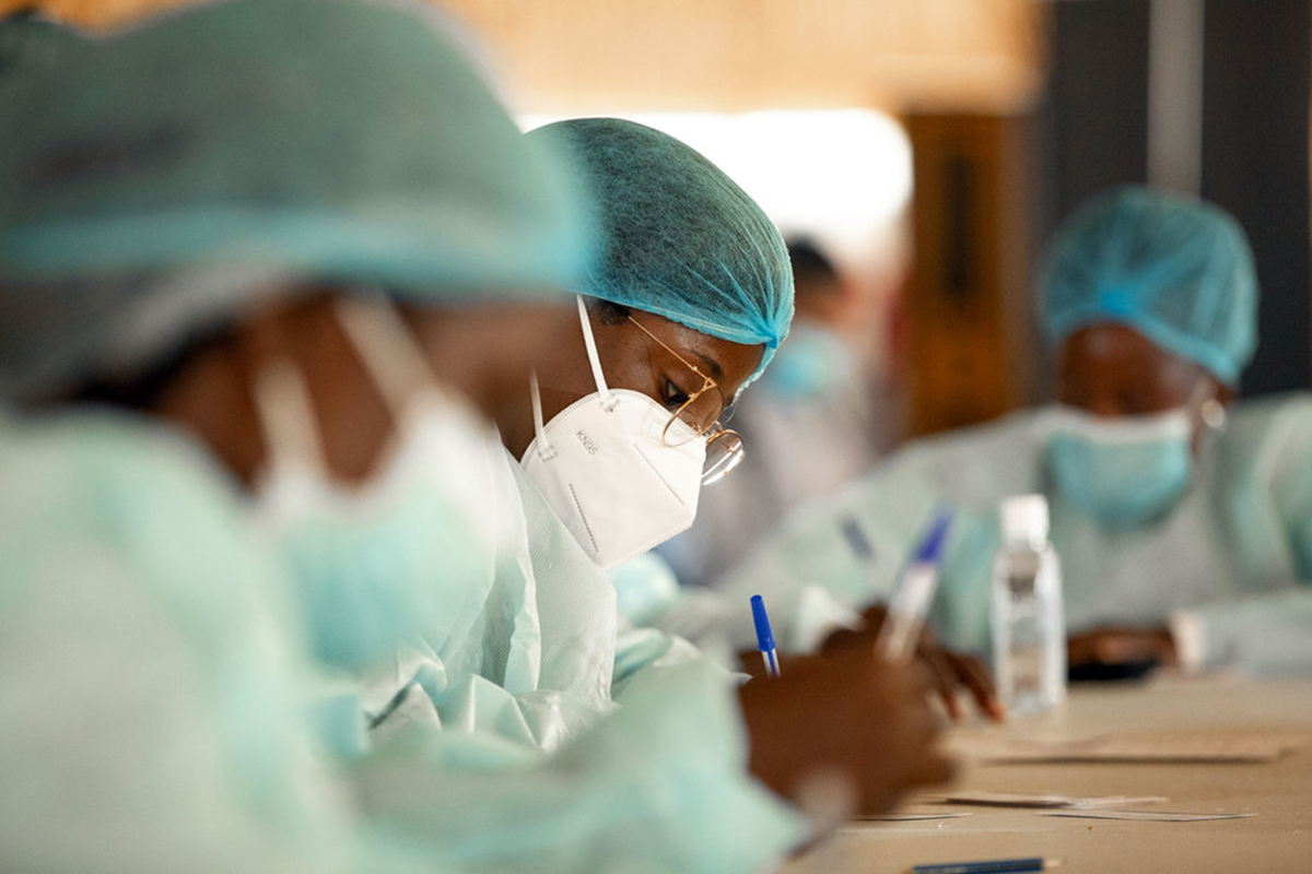 Women wearing protective gear sit at a table writing with pens