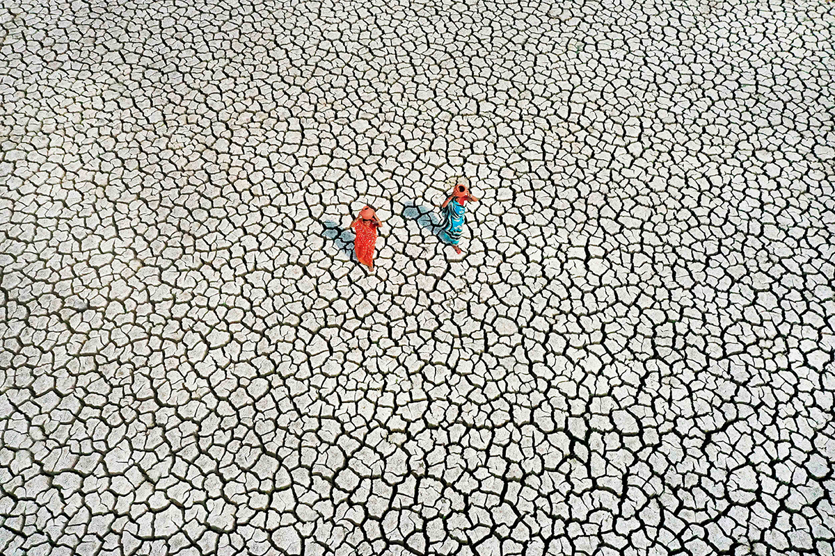 A view from above of two women walking, carrying containers on their heads, through very cracked soil.