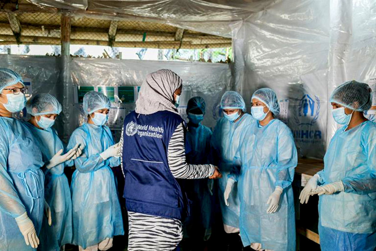 a group of health workers in protective gear gather around a woman wearing a who vest.