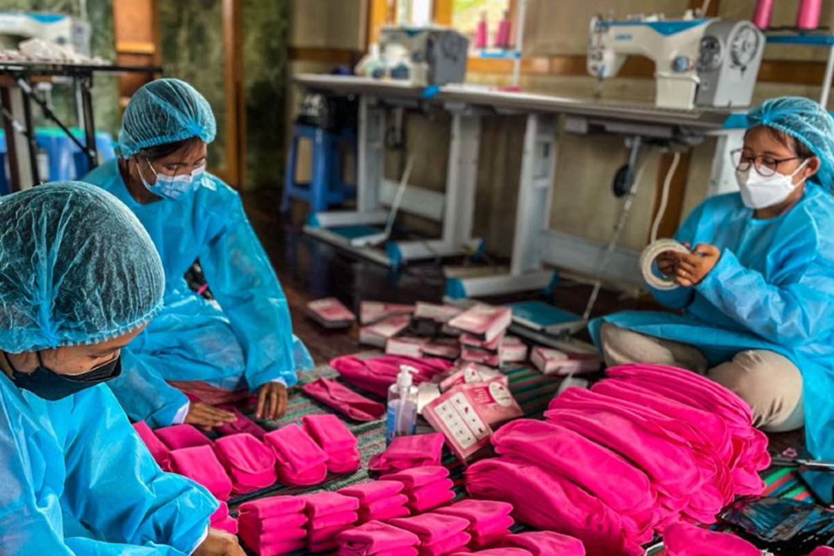 Three women sit on the floor around a mound of folded, bright pink, cloth pads.