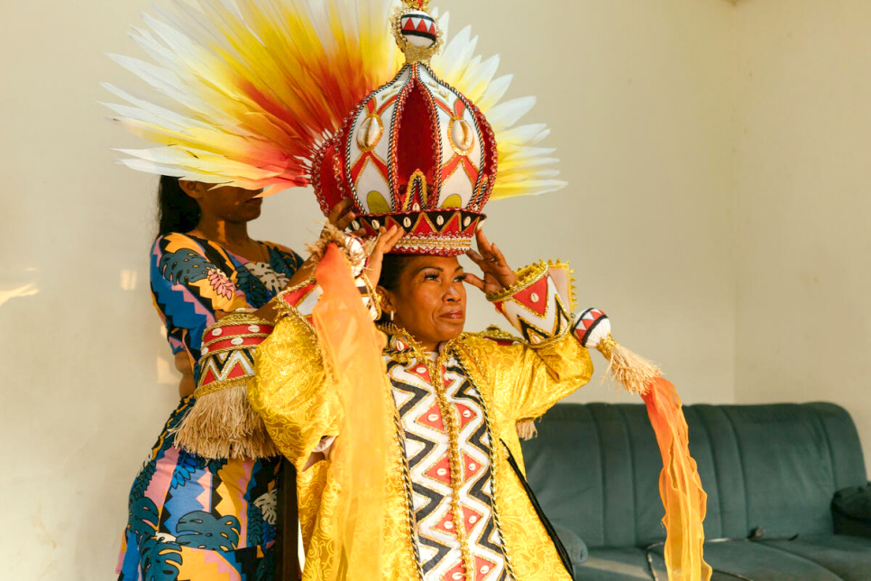 Ingrid sits in costume while another woman helps her put on a large and colourful headpiece