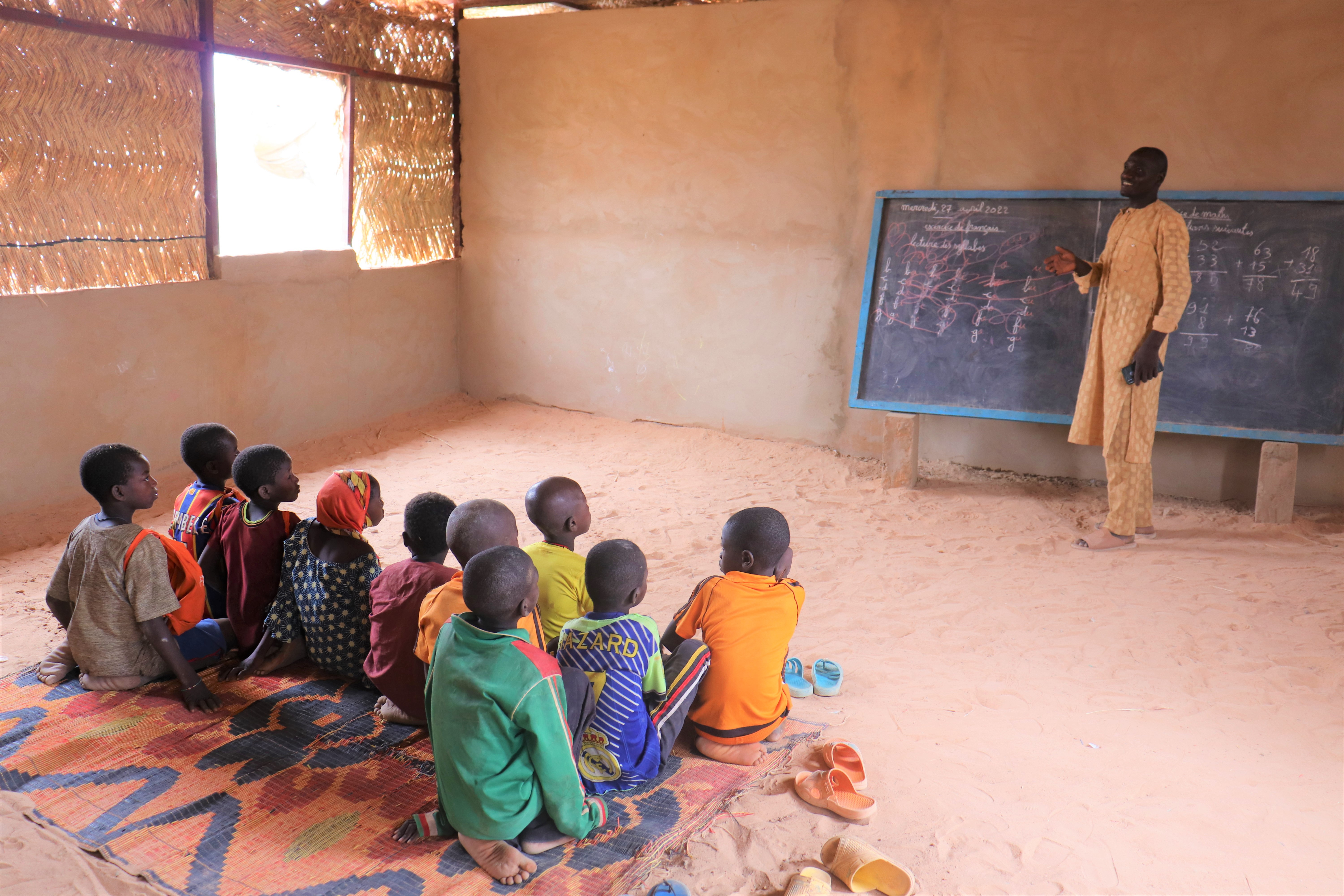 Children sit on a dirt floor inside a classroom in front of the teacher standing next to the chalkboard