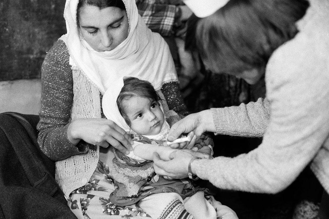 A black and white image of a girl getting a vaccine in her arm while sitting on her mom’s lap.