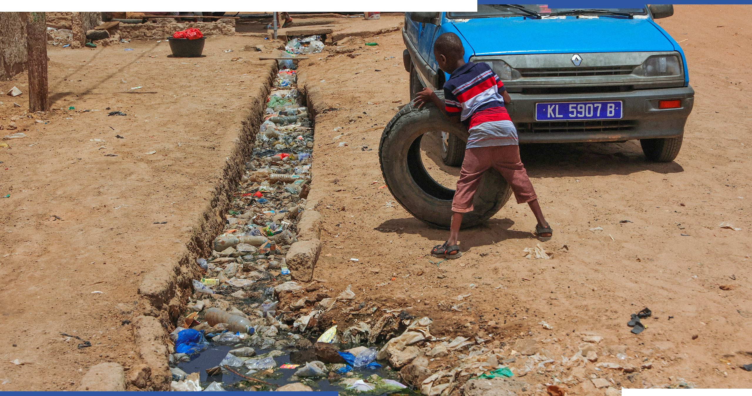 A child plays with a truck tire next to a ditch full of plastic waste