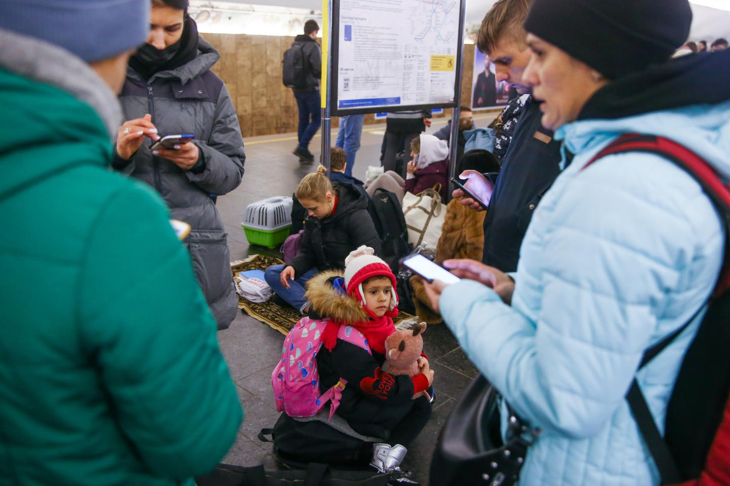 a boy with a teddy bear sits on the ground surrounded by bags and people on their phones