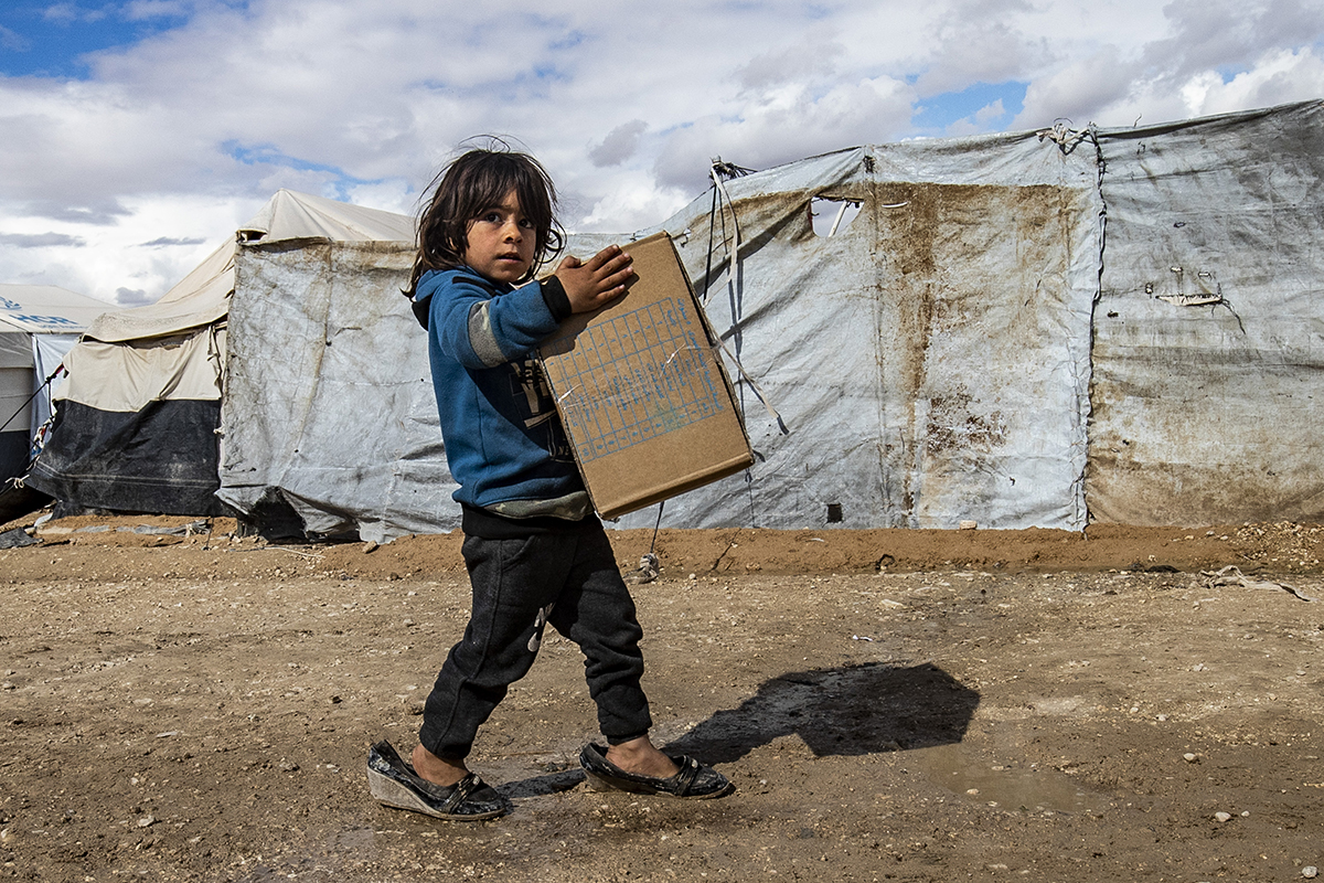 A boy wearing his mother’s shoes carries a box. 