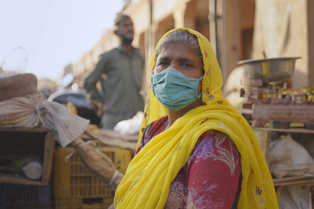 a woman wearing a facemask stands in front of harvest crates and a scale.