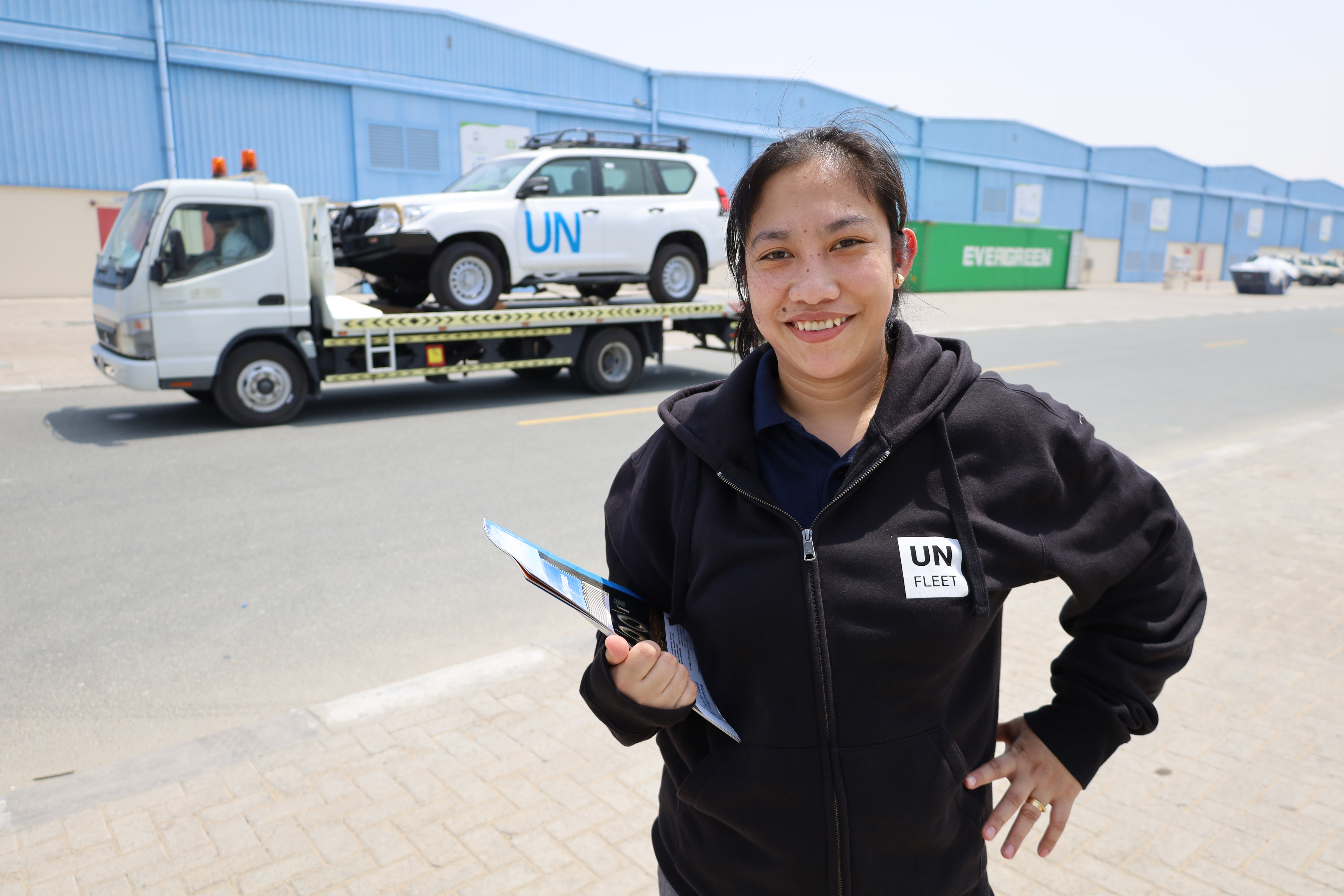 A woman stands in front of a tow truck carrying a white vehicle that reads UN in blue.