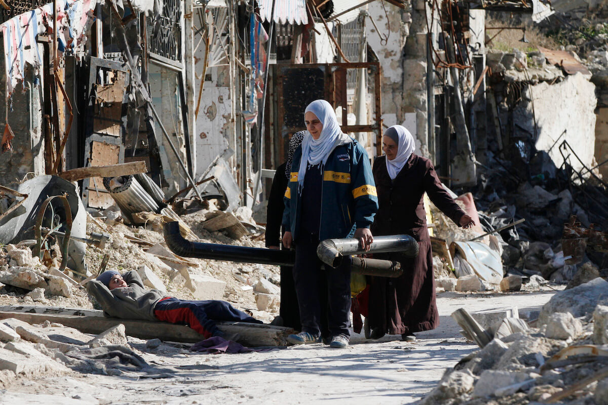 women walk carrying pipes among the rubble