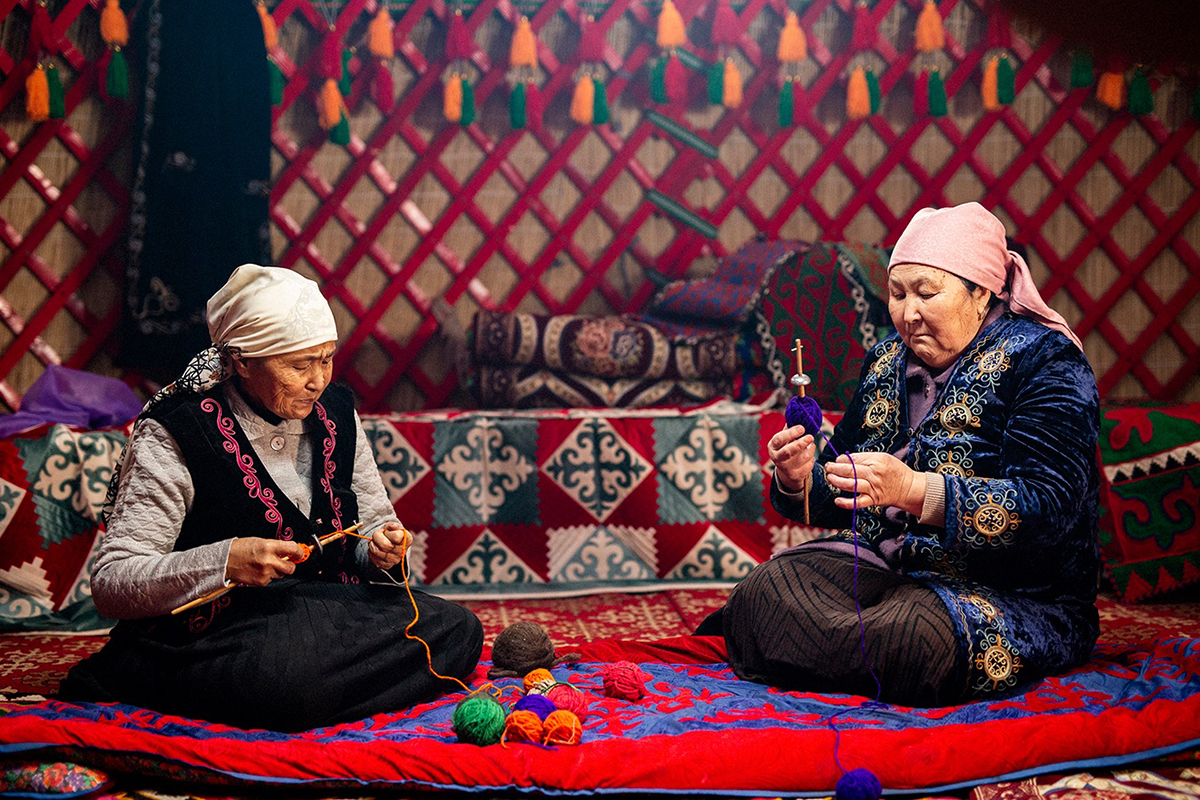Two women sit on a rug spinning wool yarn.