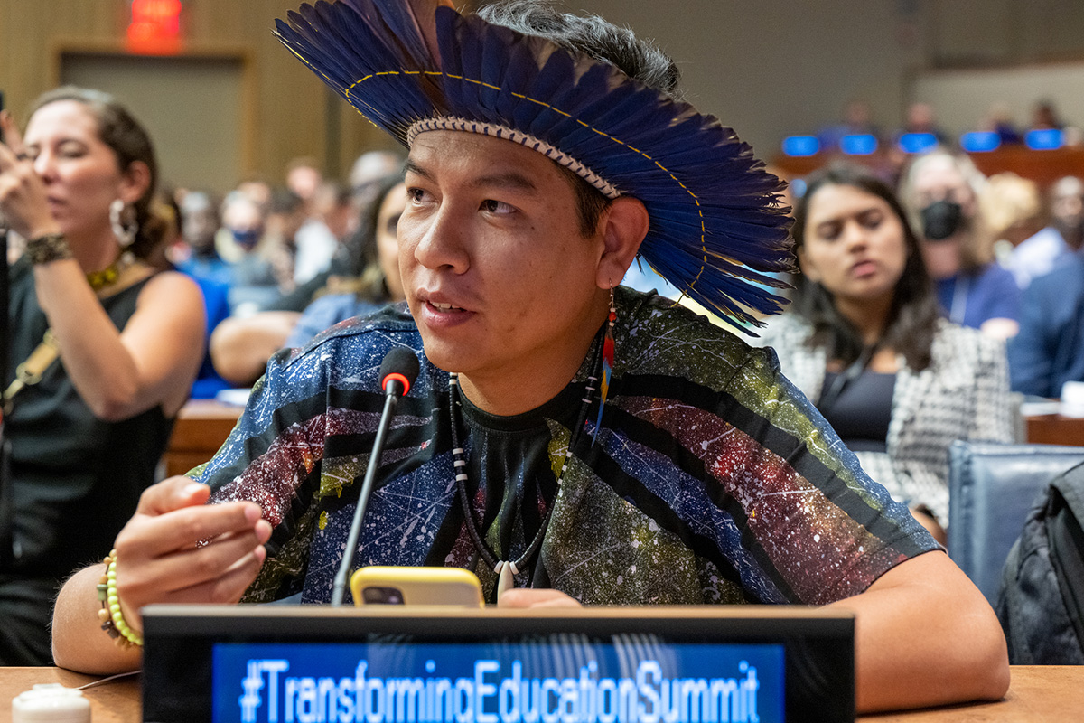 An indigenous youth leader in traditional attire speaks into a microphone.