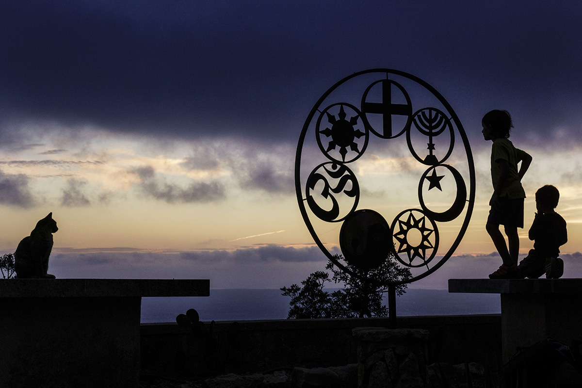 a metal sculpture with different religious symbols with kids playing around it at dusk