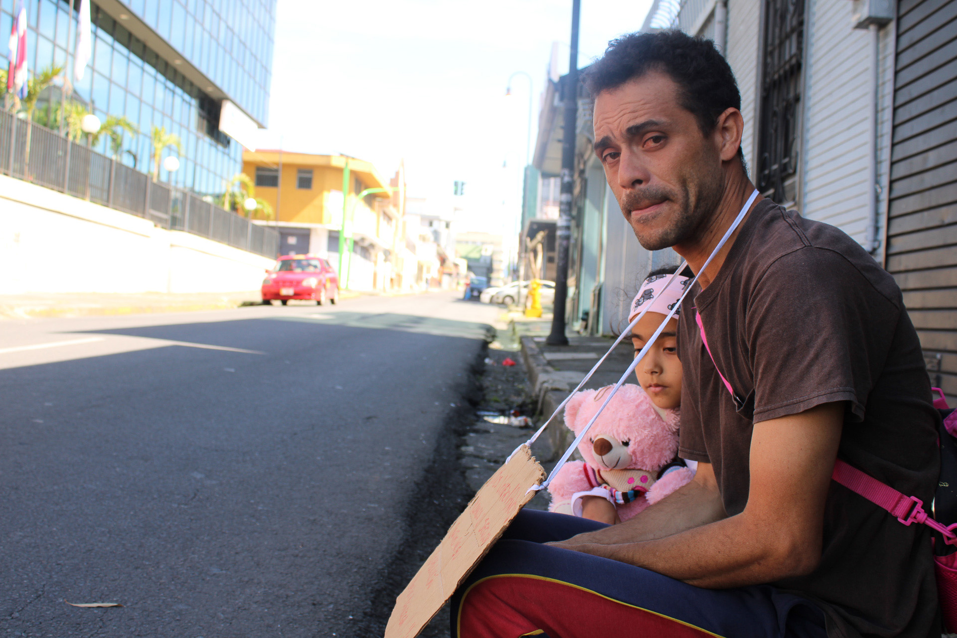 A man and his daughter sit in the street holding a cardboard sign.