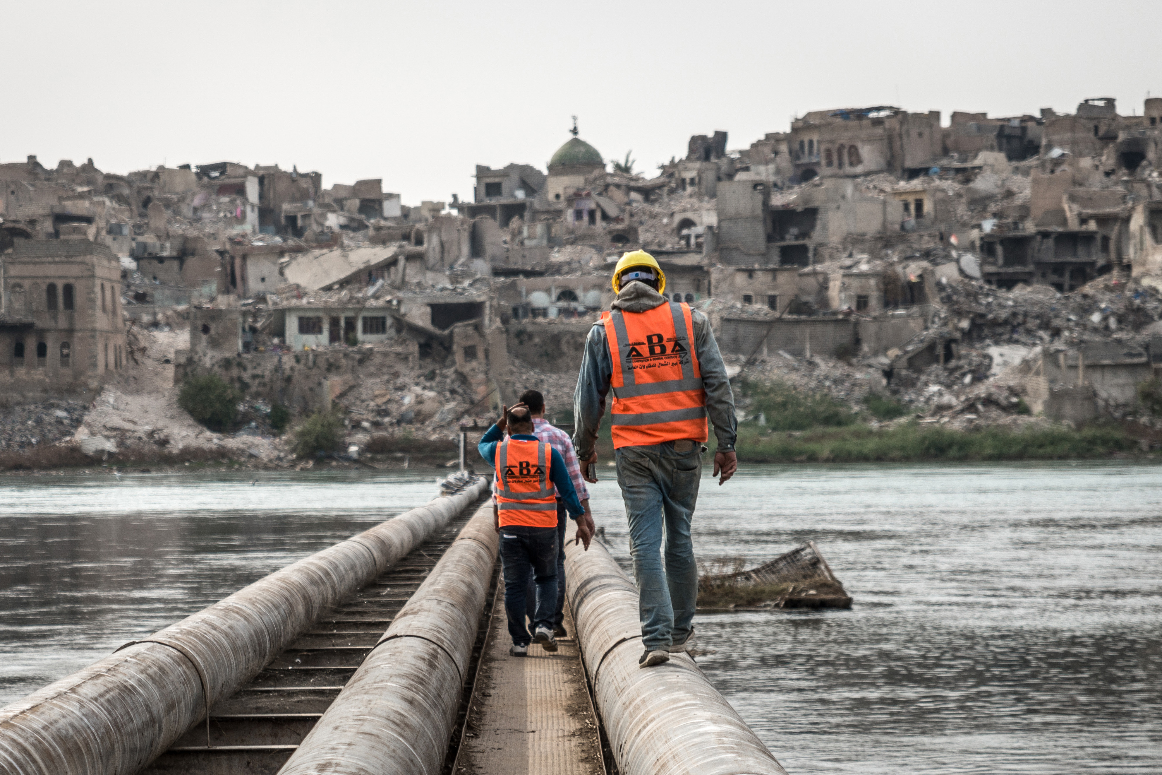 Three men walking on pipes across a river towards a town in ruins