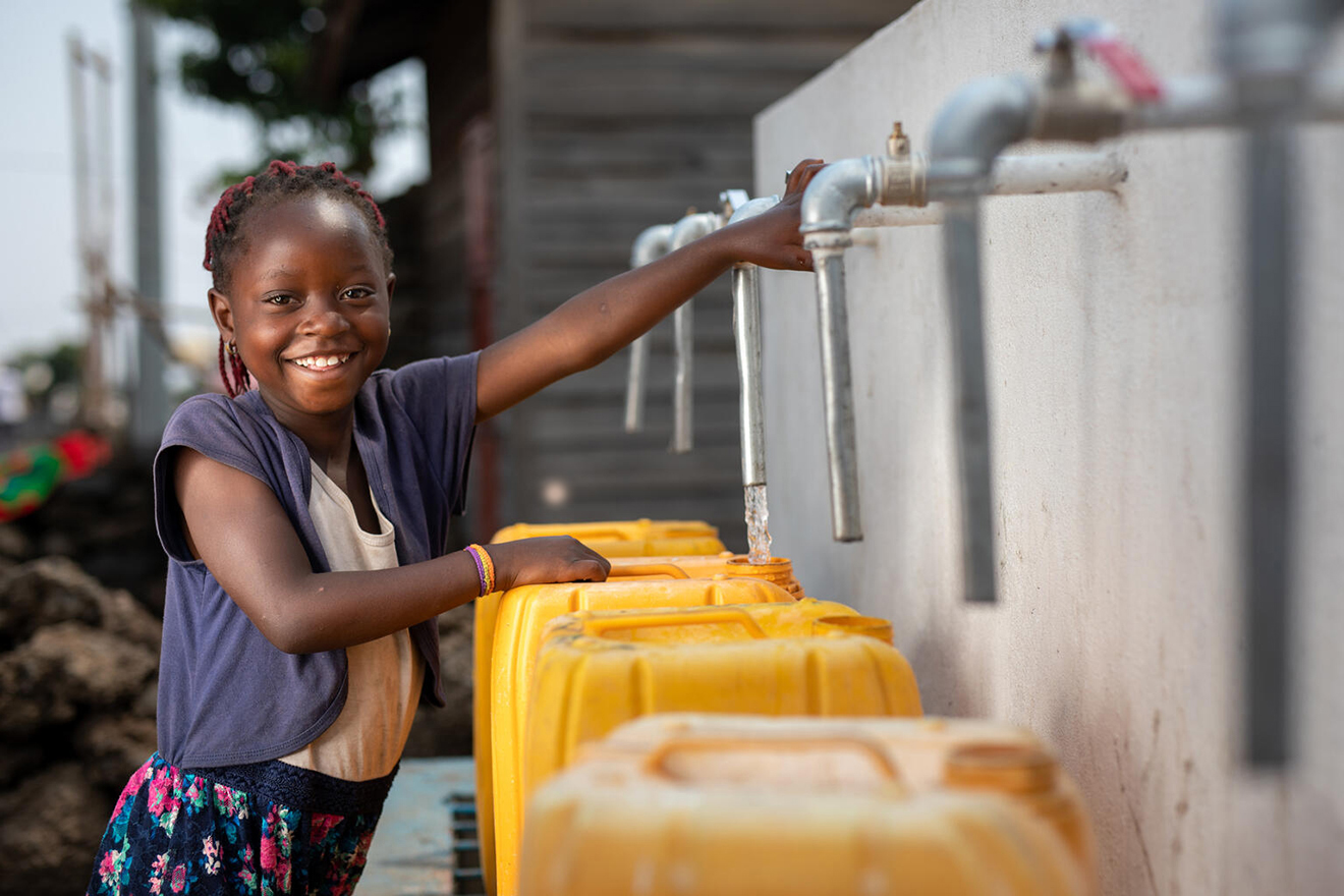 A smiling girl draws water from a standpipe