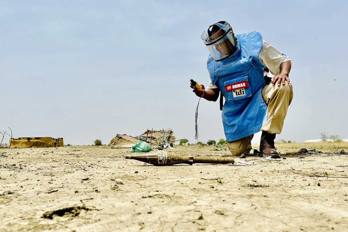 A man in protective gear kneels in front of a landmine while holding a device.