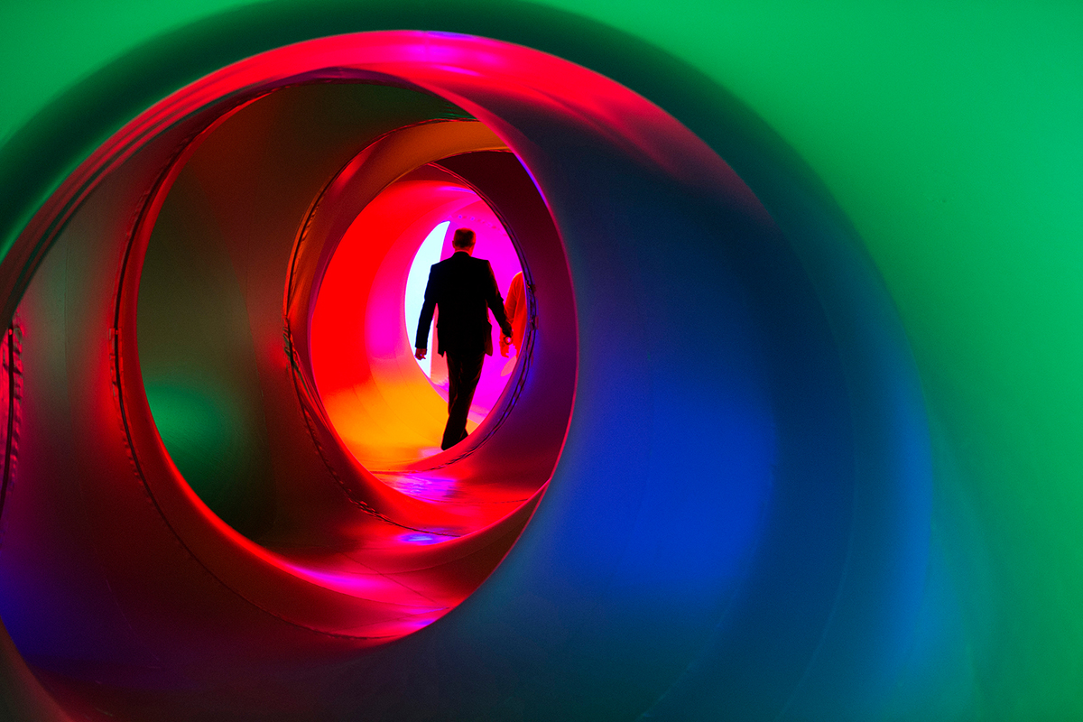 The silhouette of a person at the end of a round winding corridor illuminated by multi-coloured light.