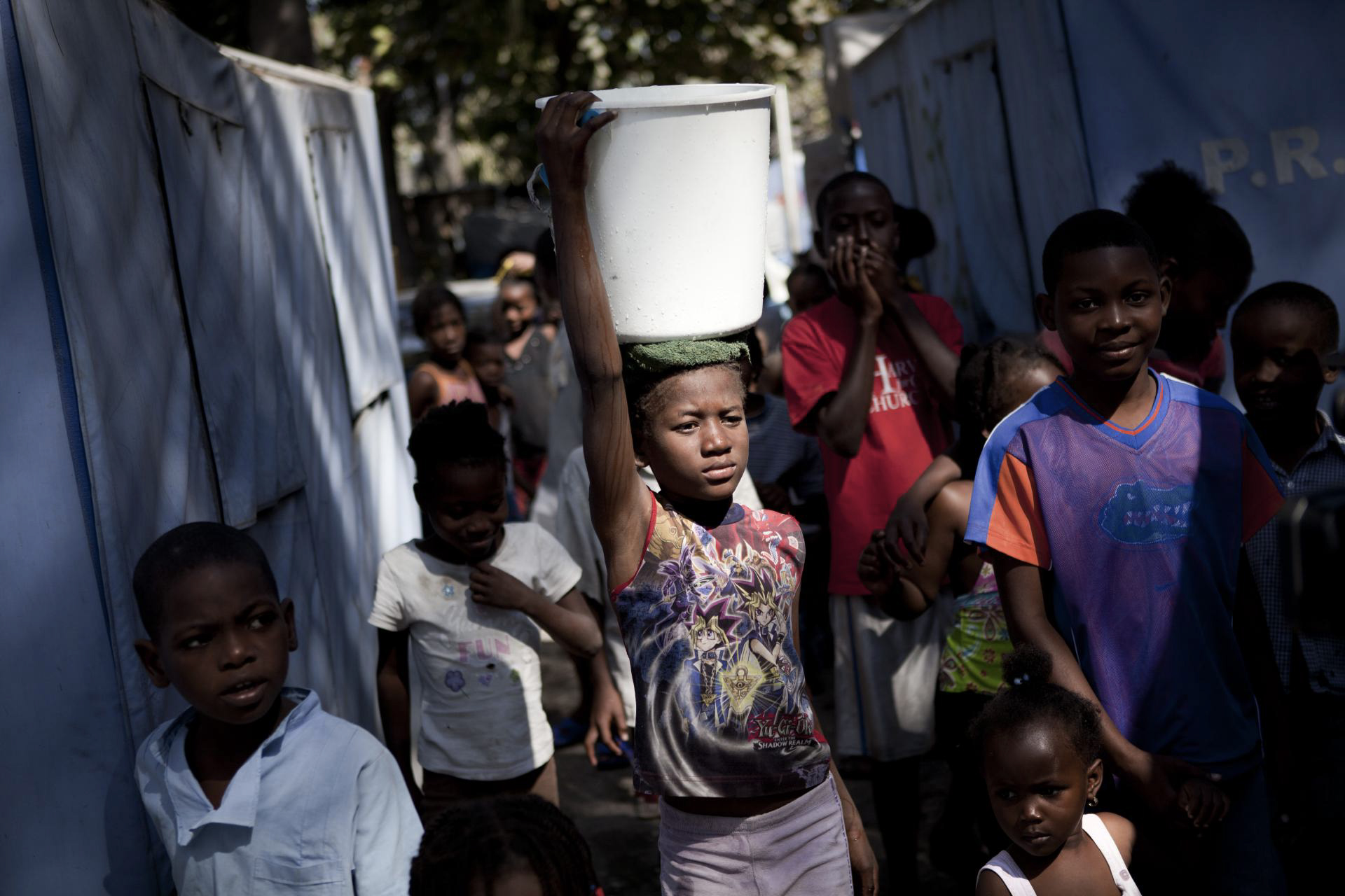 A girl carries a water bucket on her head among a crowd.