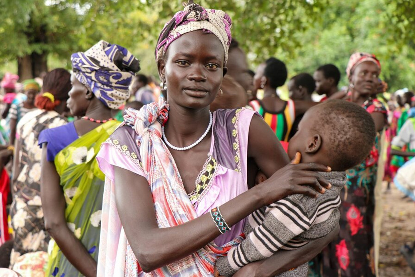 A woman looks at a camera while holding her kid surrounded by other women. 
