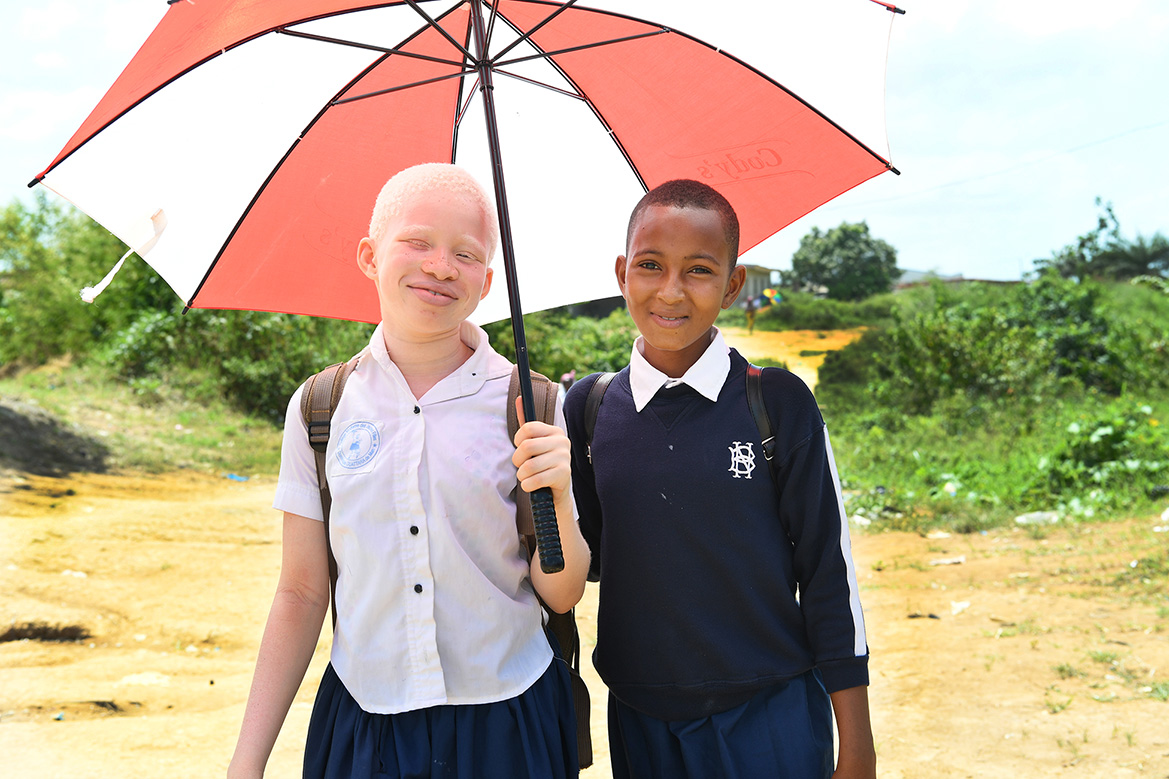Two girls in uniform smile and share an umbrella to cover from the sun