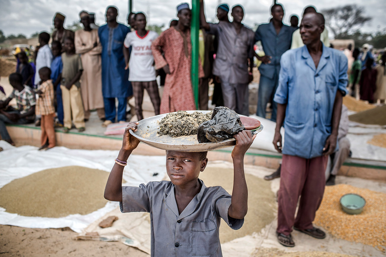 A boy carries some goods on his head at a local market and people wait in the background