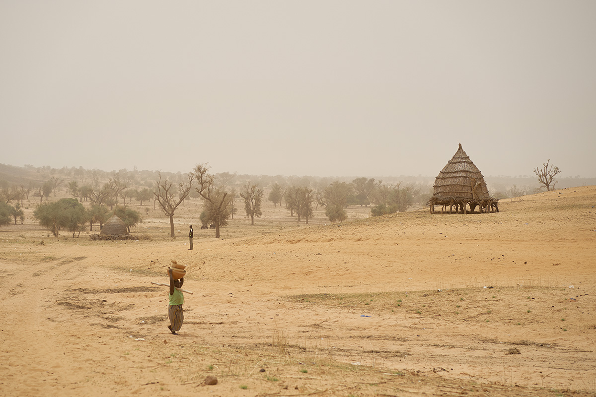 In a vary arid landscape with two round straw huts, two persons stand about.