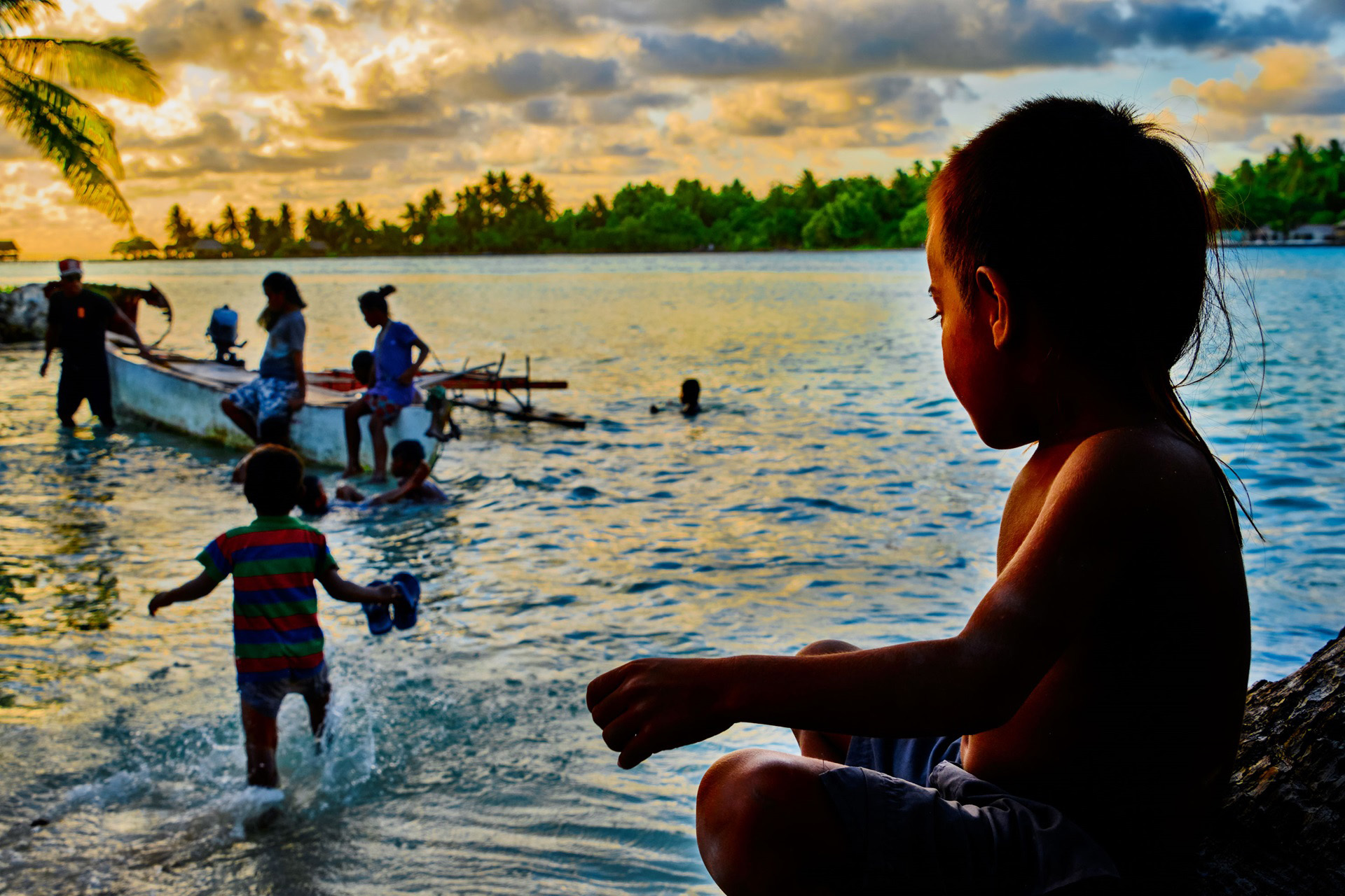 a boy looks on from shore at people on and swimming around a boat