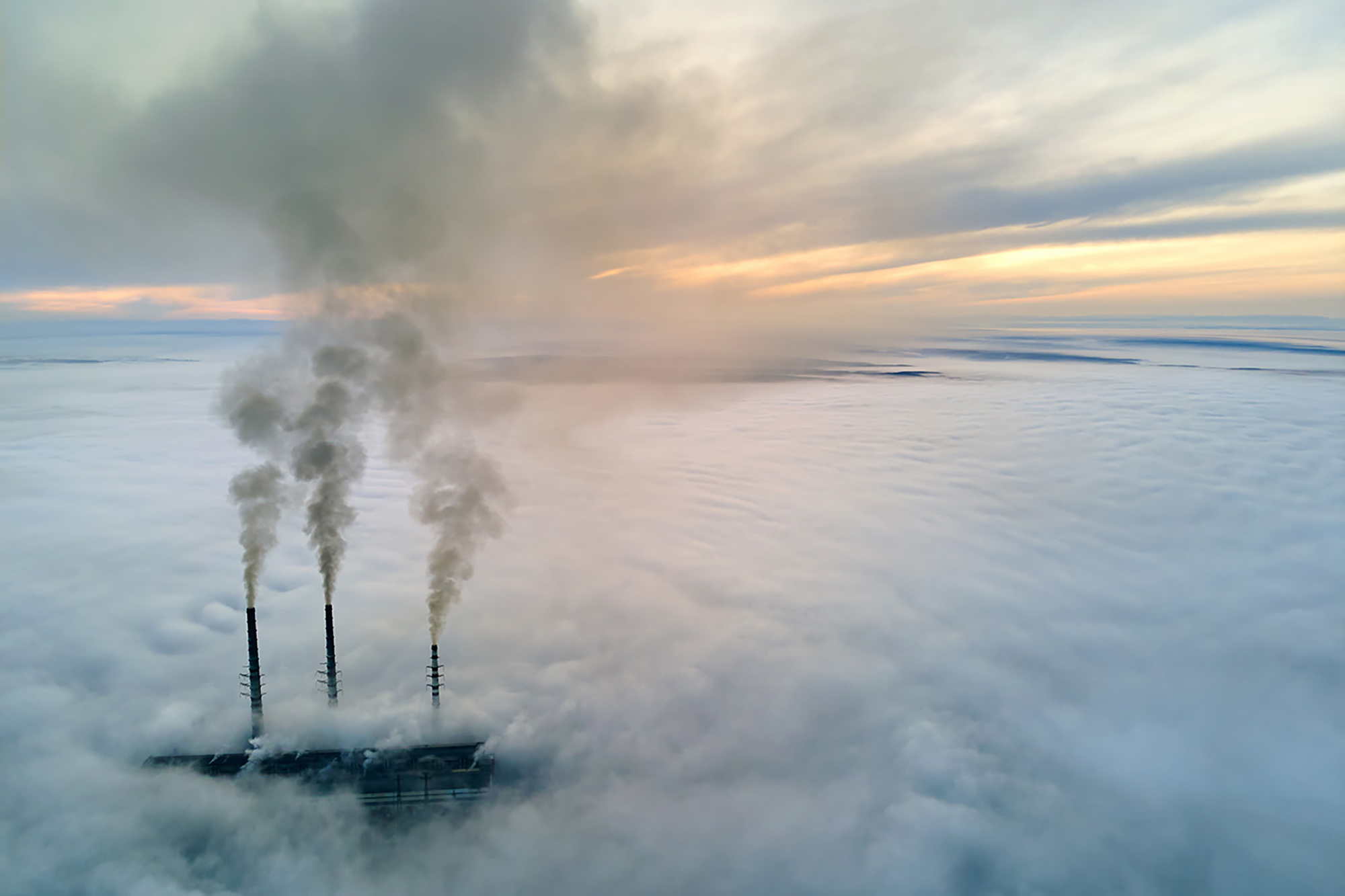 Aerial view of a coal-fired power plant and tall smokestacks rising and polluting the atmosphere as the sun goes down.