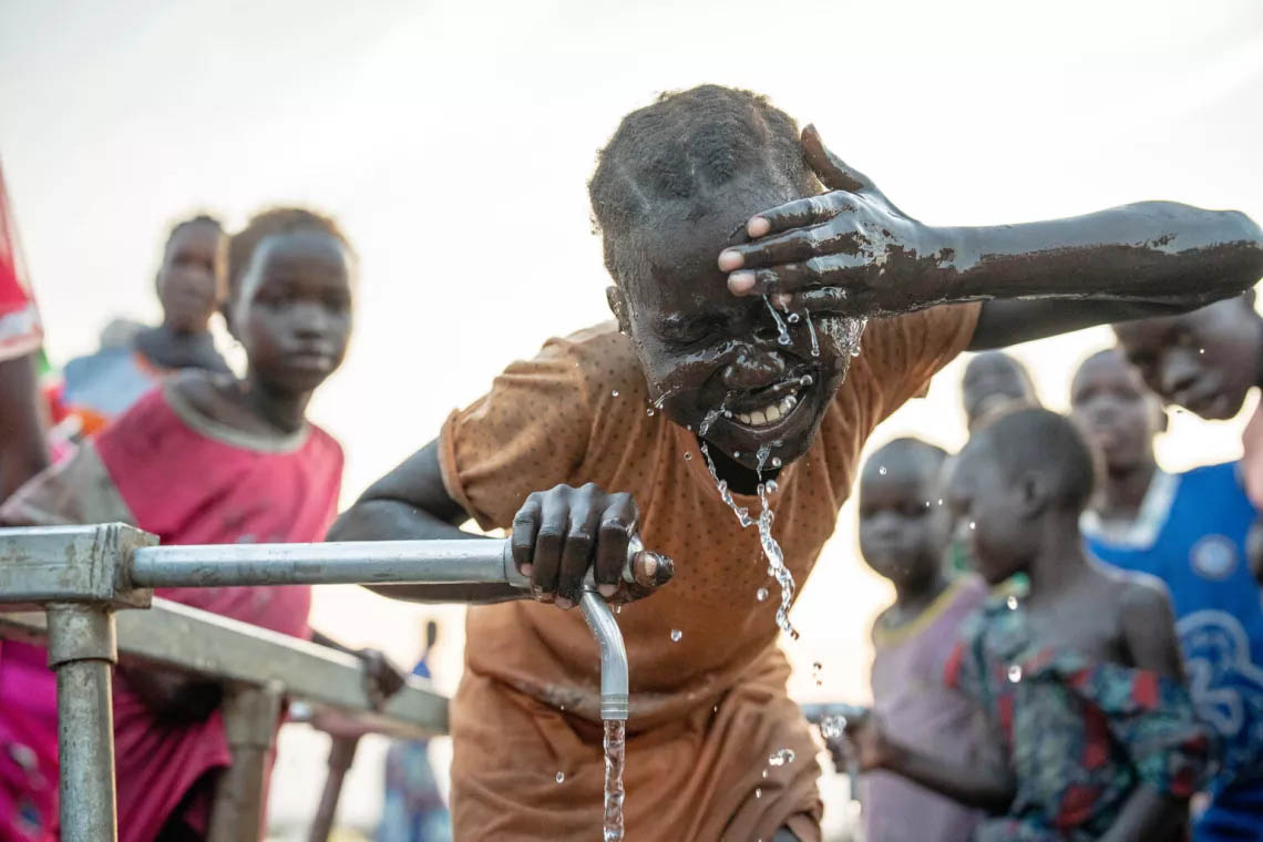 A young girl surrounded by children washing her face a well pump.