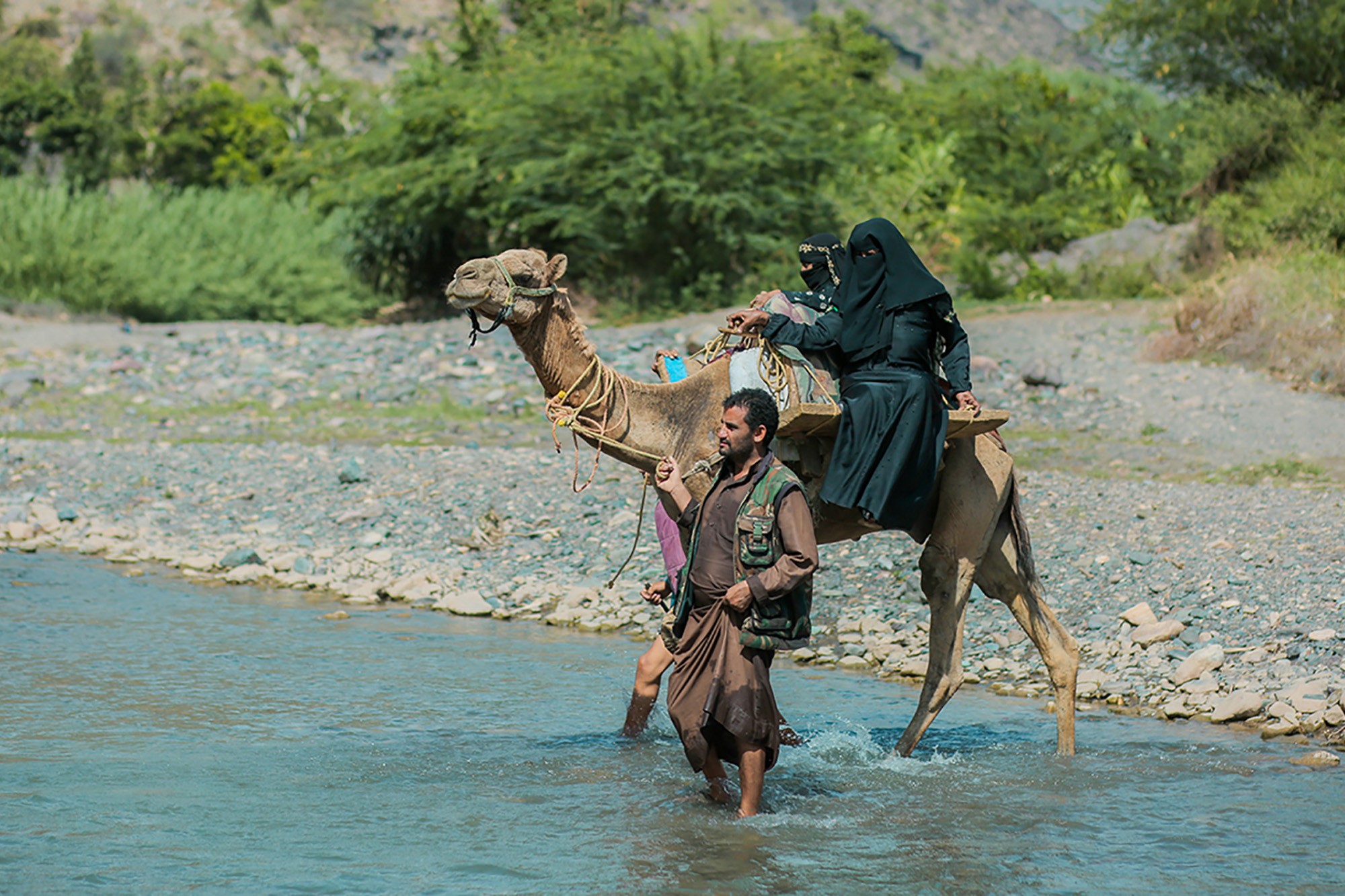 A pregnant woman is crossing a river on a camel that is being pulled by a man.