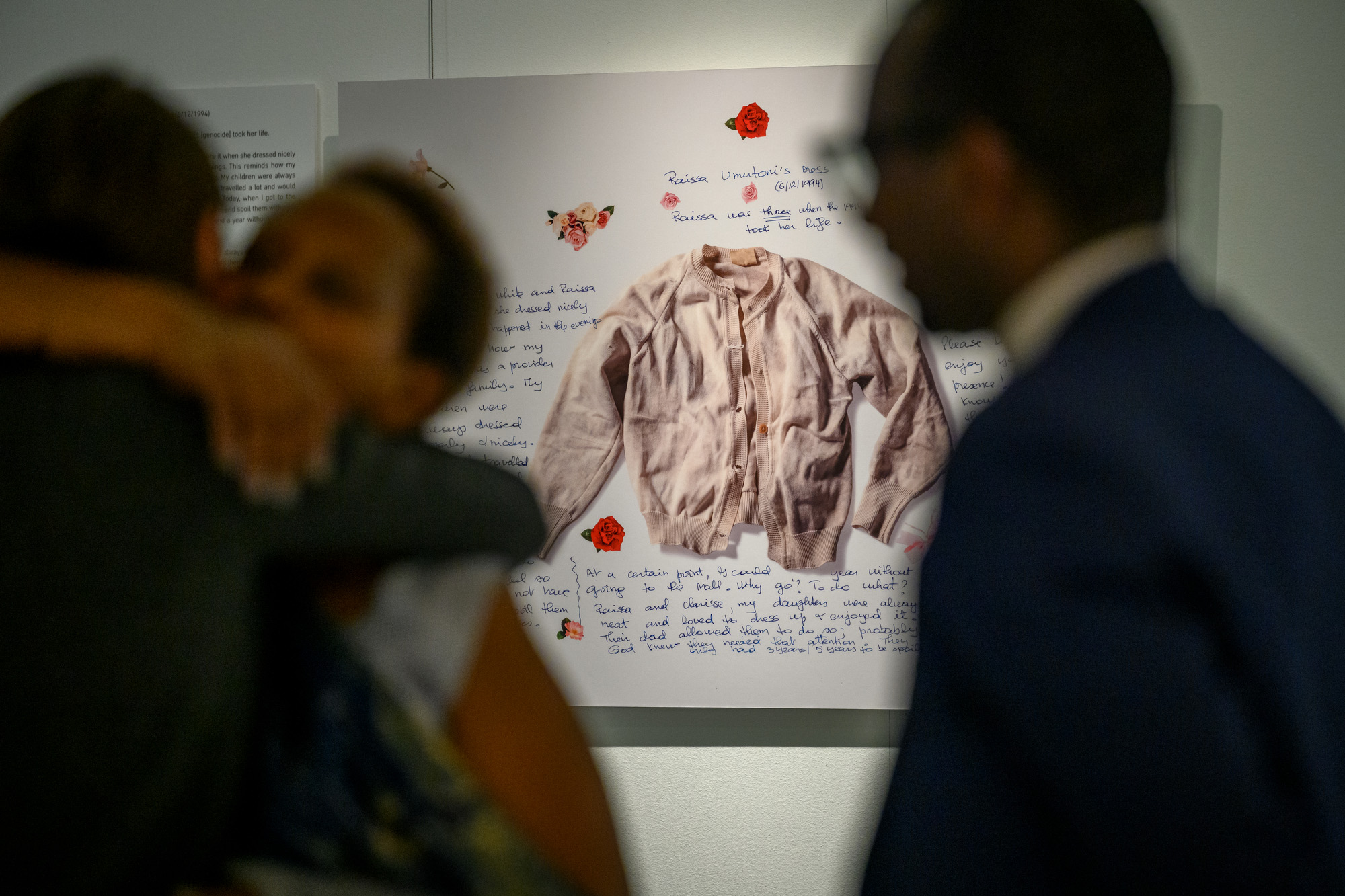As part of an exhibit on genocide prevention, a framed shirt surrounded by text is shown in the background, while two people embracing in the foreground are blurred.