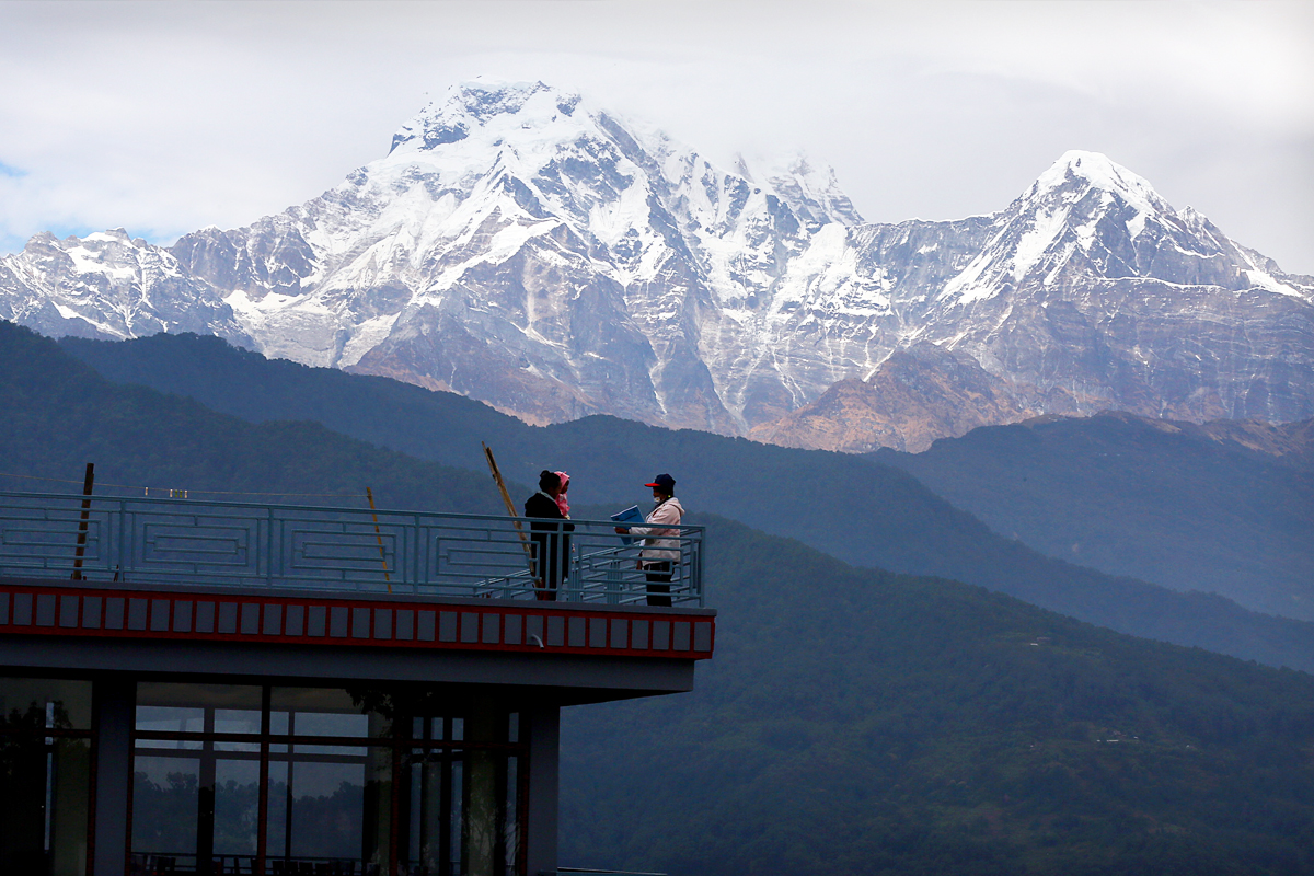 Two people stand facing each other on a rooftop with snow-capped mountains in the distance